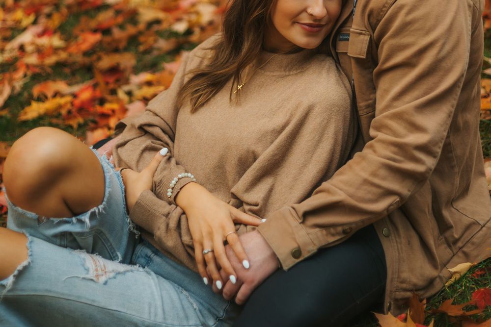 Couple sitting together on autumn leaves, woman in ripped jeans and beige sweater, man in brown jacket.
