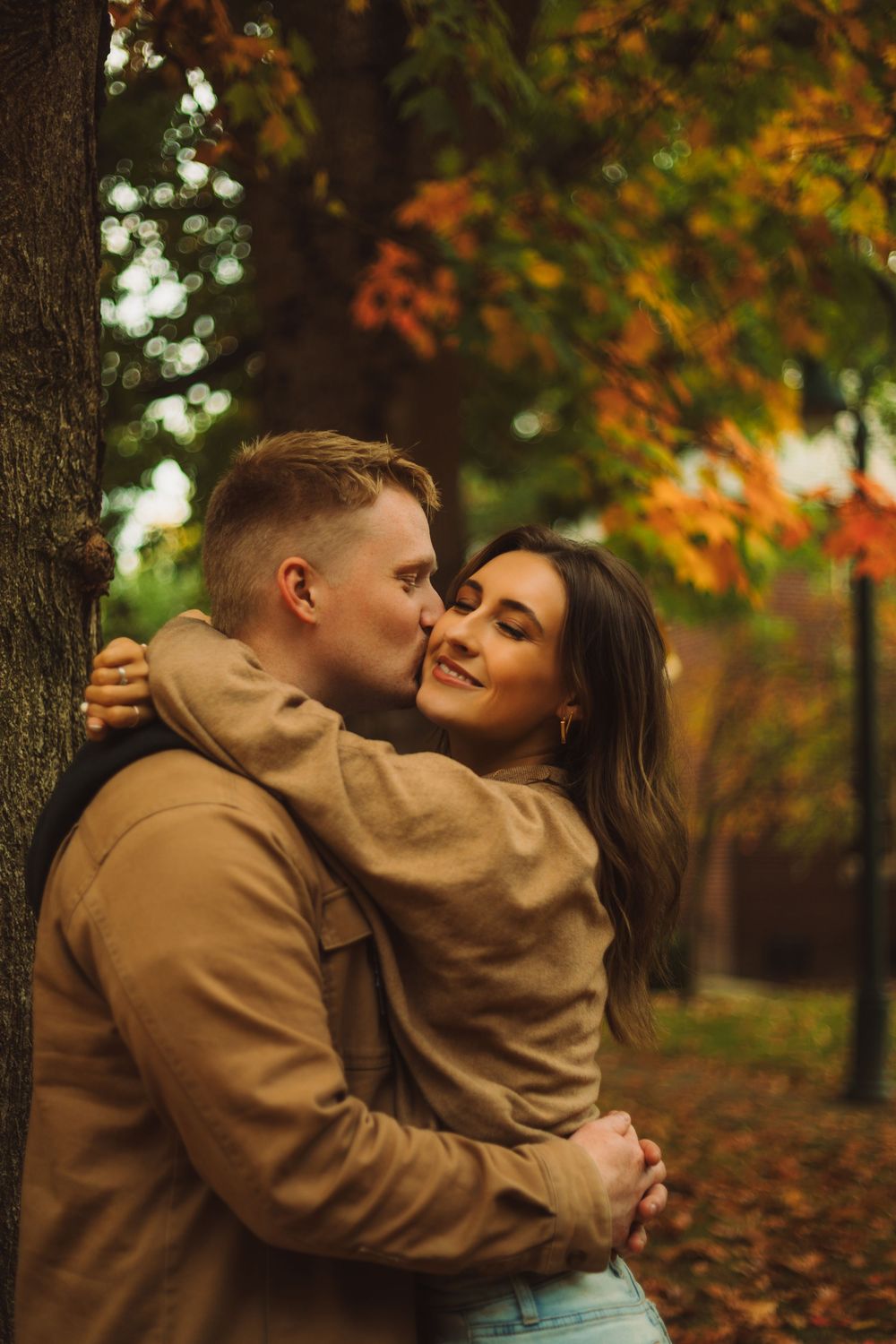 Couple embracing and kissing by a tree in an autumn park with colorful orange and yellow foliage.