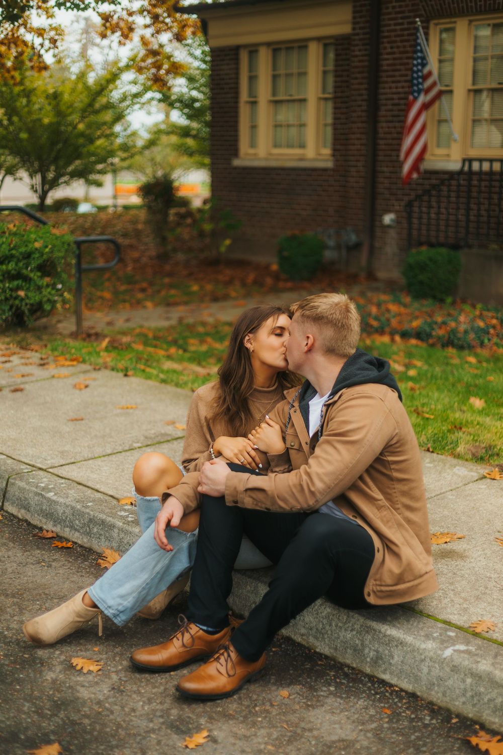 Couple sharing a kiss on front steps of brick home with American flag in autumn.