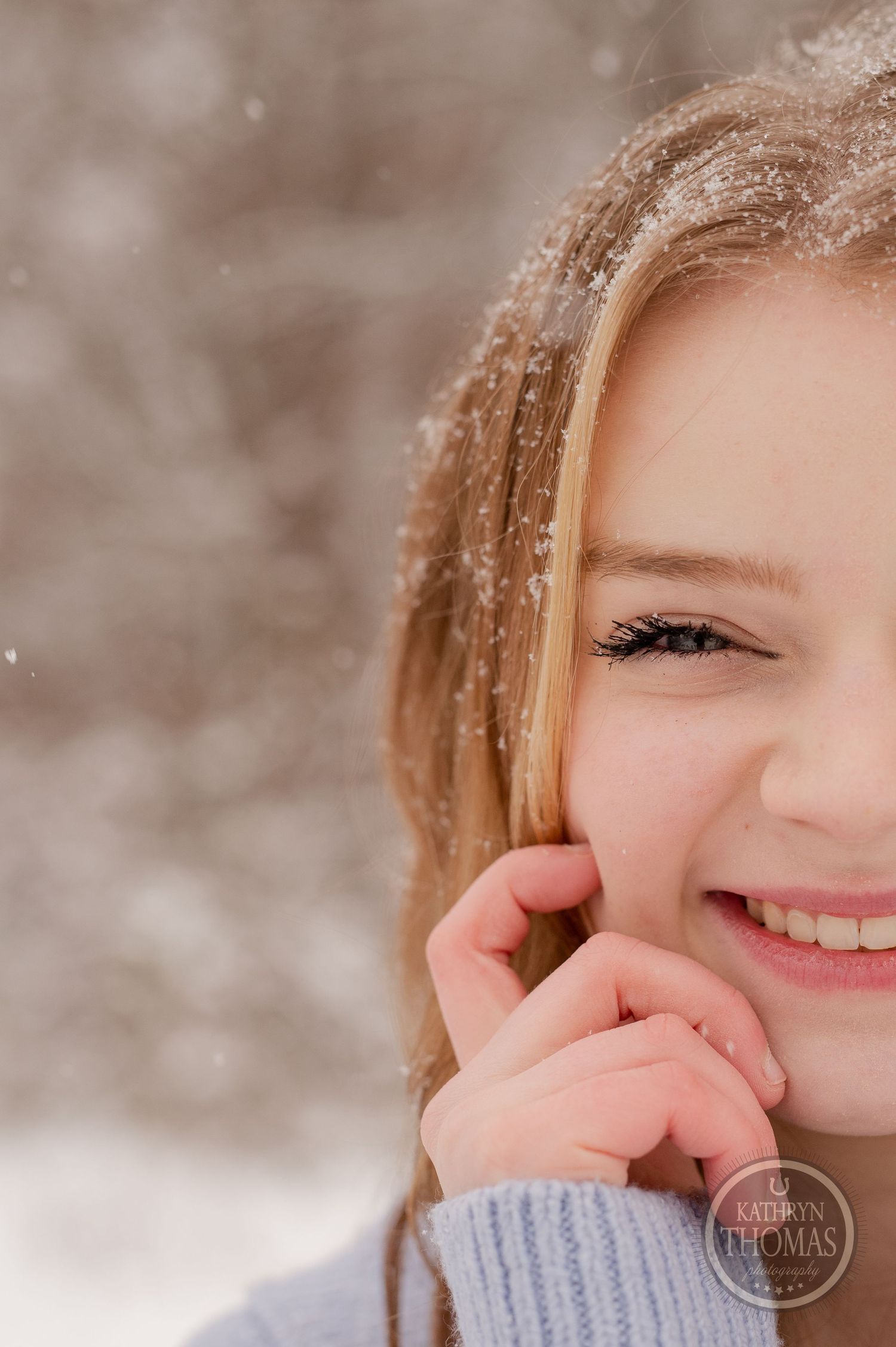 A close-up winter portrait showing a bright smile with snowflakes falling in the background.