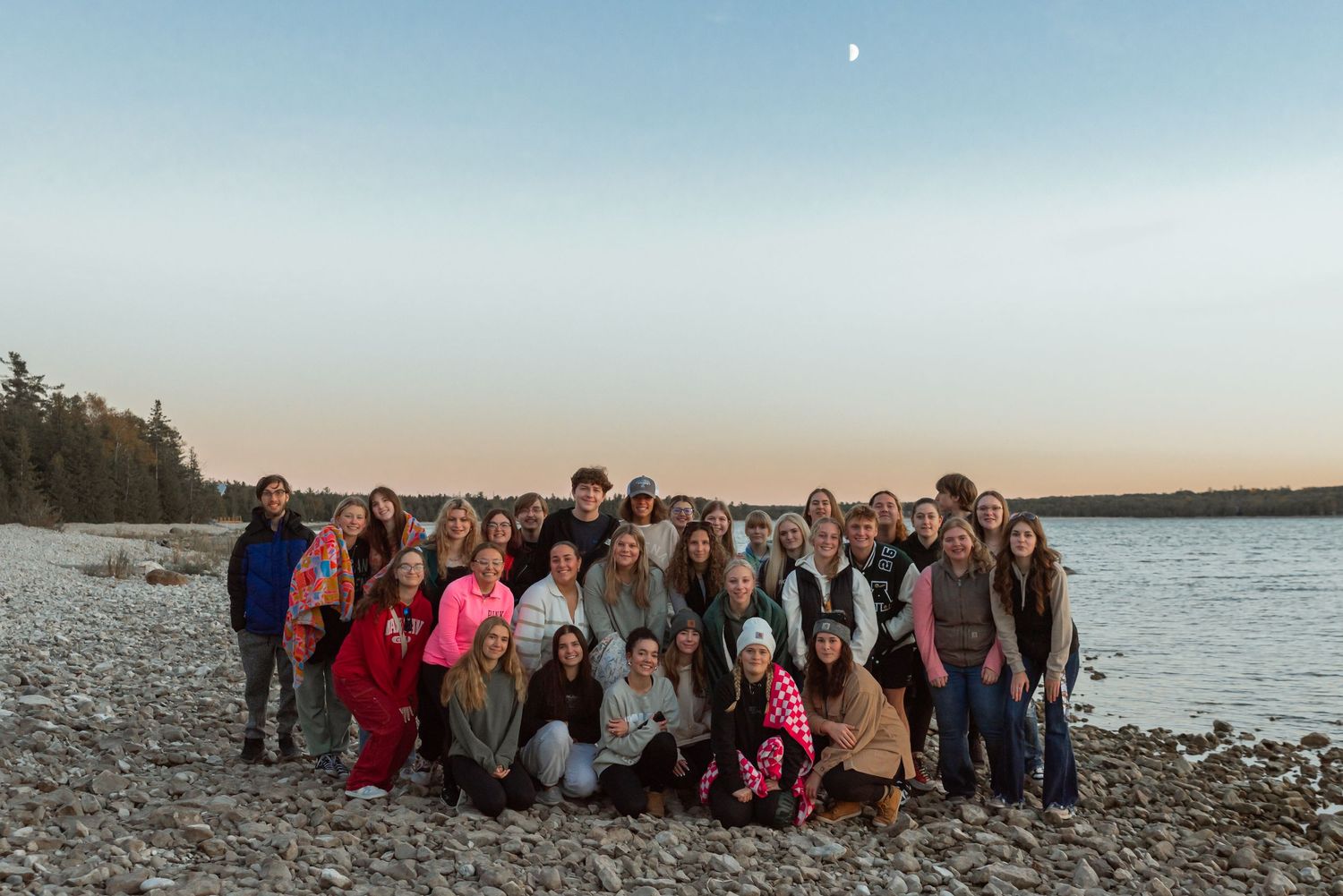 Large group photo of students gathered on a rocky beach at sunset with water and trees in background.