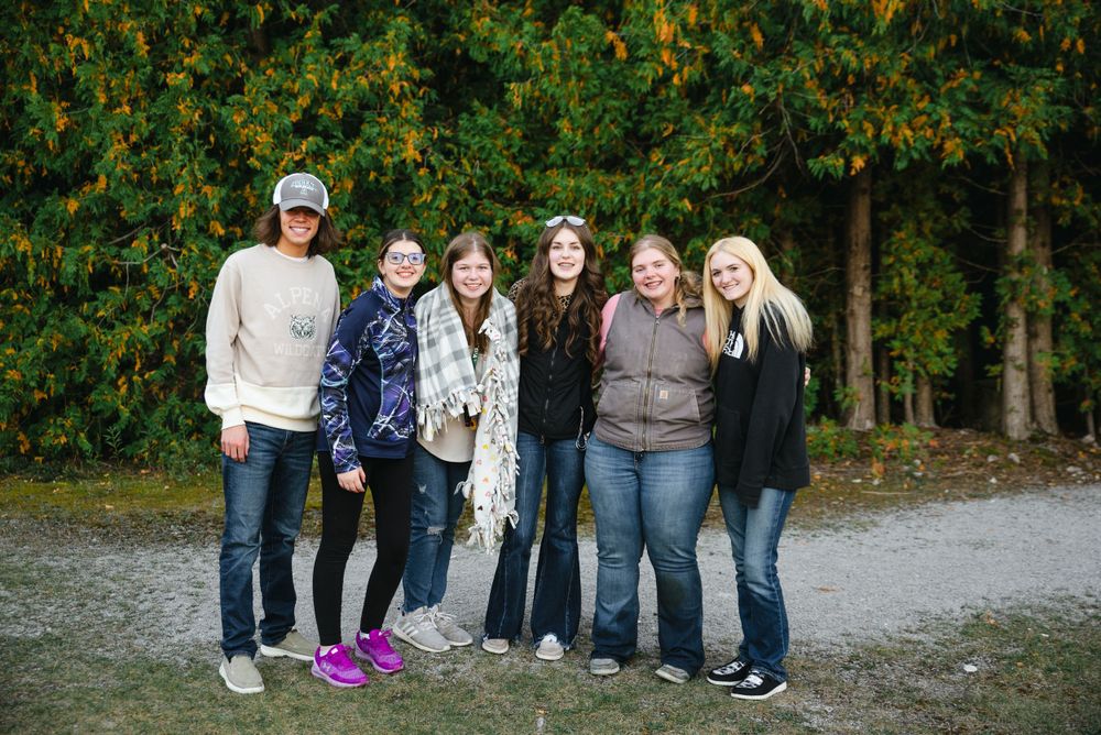 Group of six friends pose together on a fall day with colorful autumn foliage in the background.