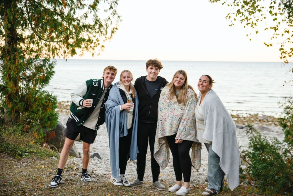 Five friends wrapped in blankets stand together on a beach path at sunset.