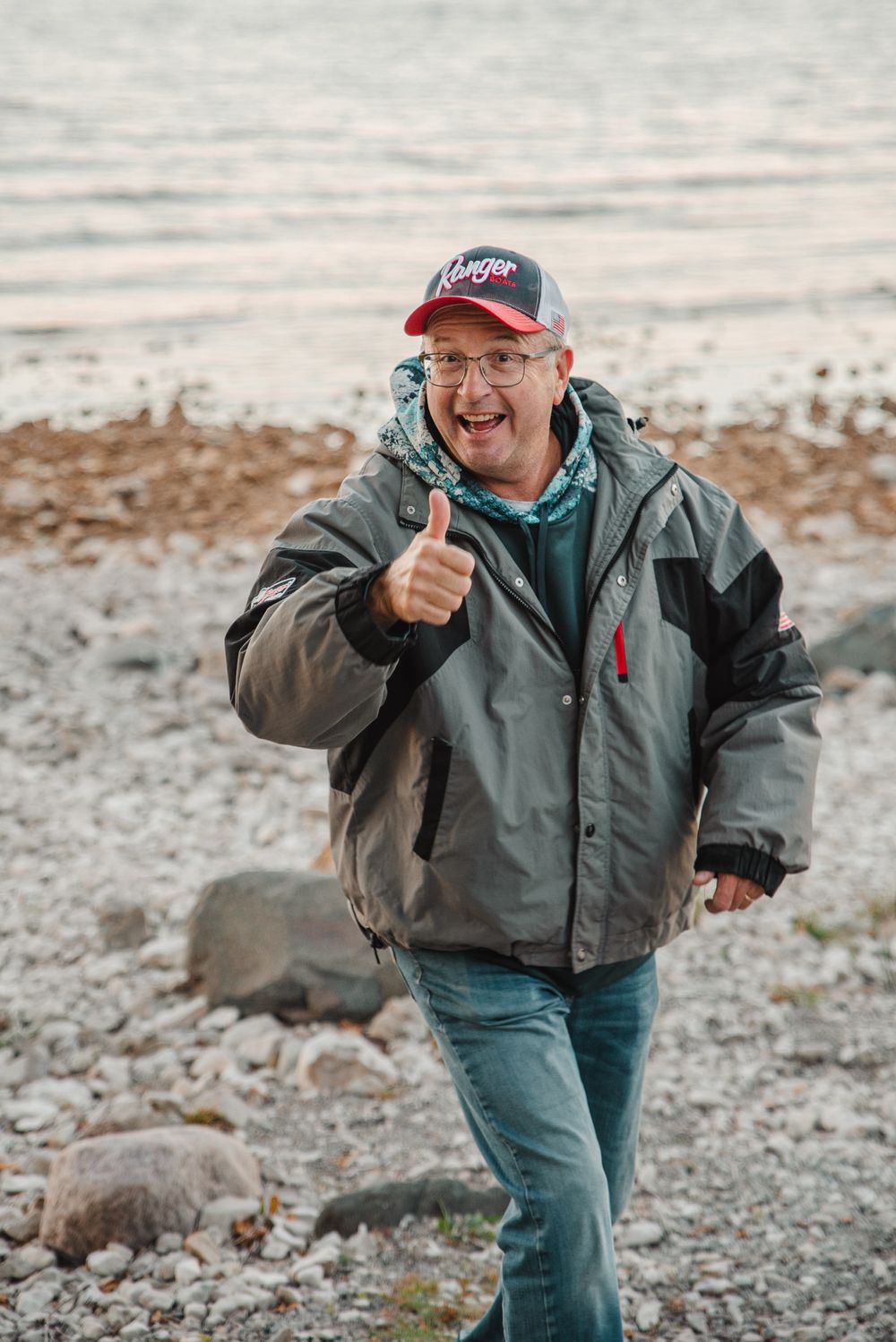Person in outdoor gear gives thumbs up while standing on rocky beach shore.