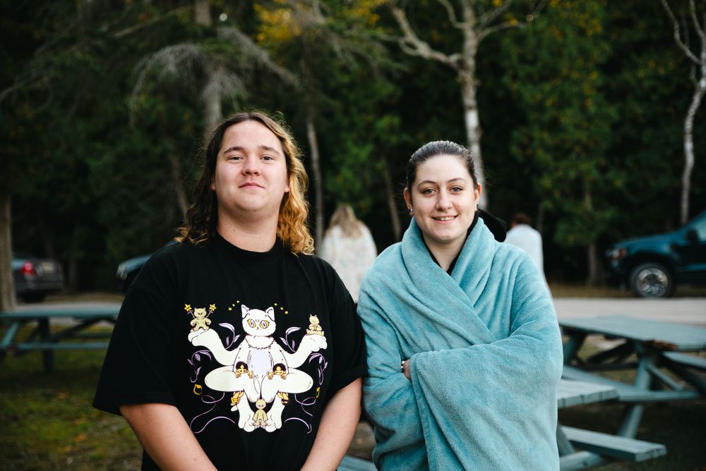 Two people sitting outdoors wearing casual clothing, one in a black t-shirt with graphic print and one in a teal wrap.