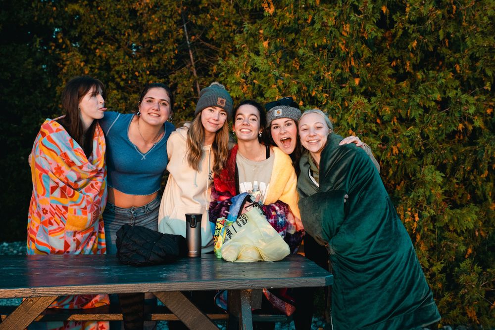Group of friends posing together on a picnic bench surrounded by fall foliage at dusk.