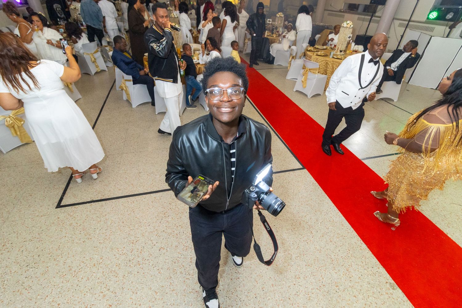 A photographer in a black jacket and blue glasses smiles while working at an event with a red carpet.