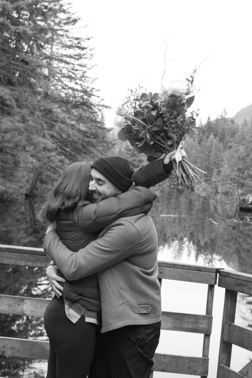 Two people embrace on a wooden deck overlooking a serene winter lake surrounded by pine trees.