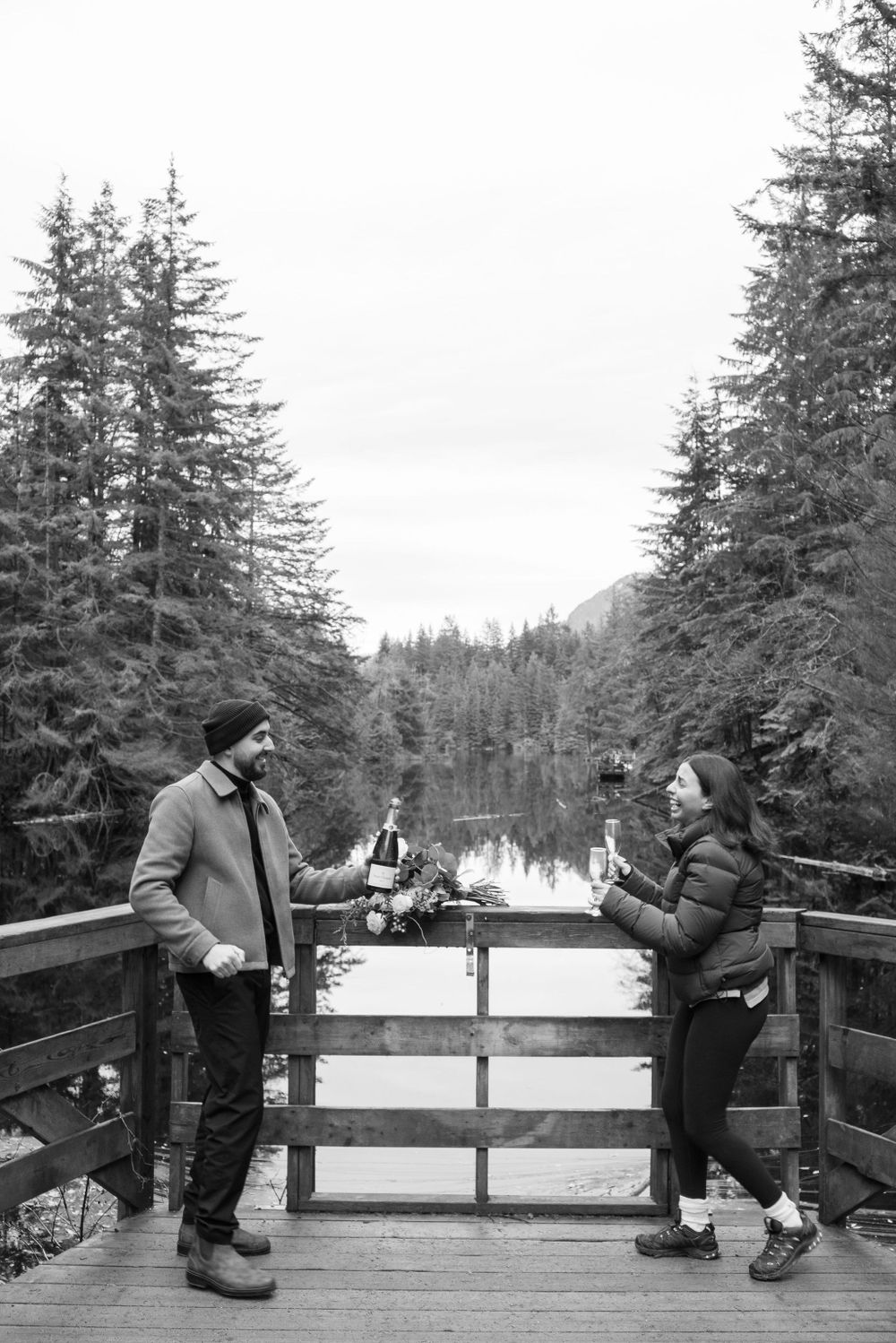 Two people share a playful moment on a wooden observation deck with mountain lake views.