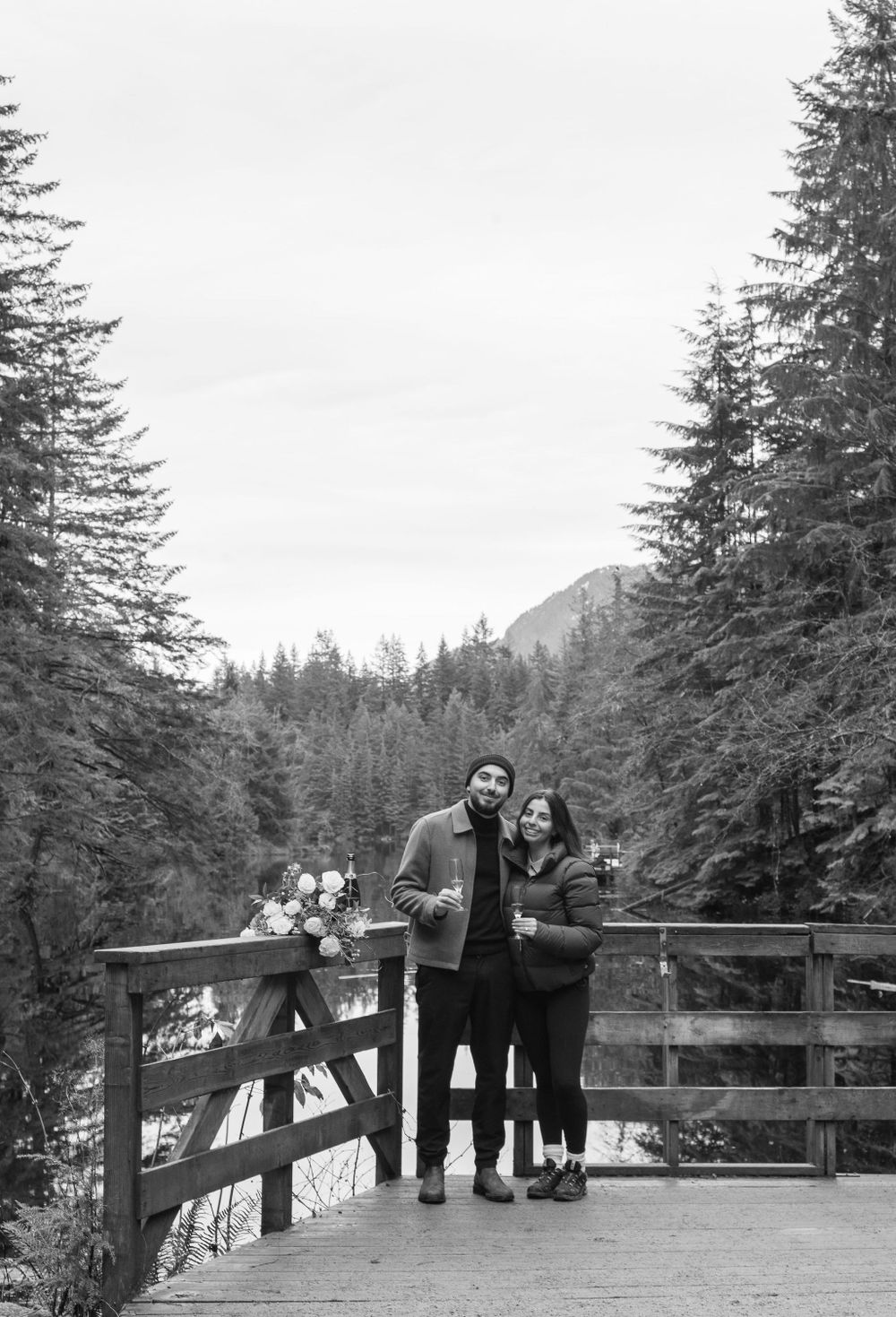 A couple stands together on a wooden observation deck overlooking a snow-dusted mountain lake.