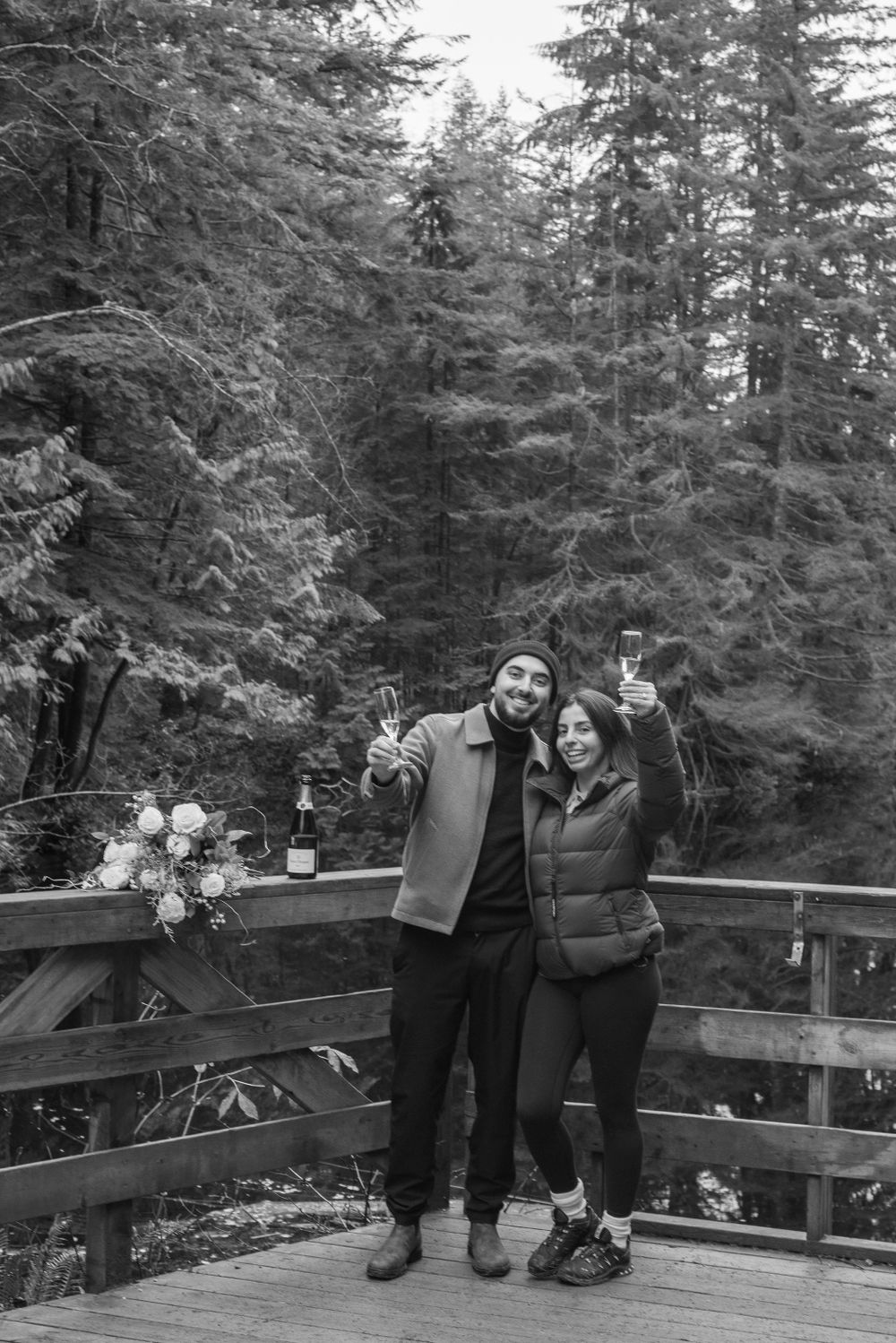 Two figures sitting on a wooden deck surrounded by evergreen trees in a black and white photo.