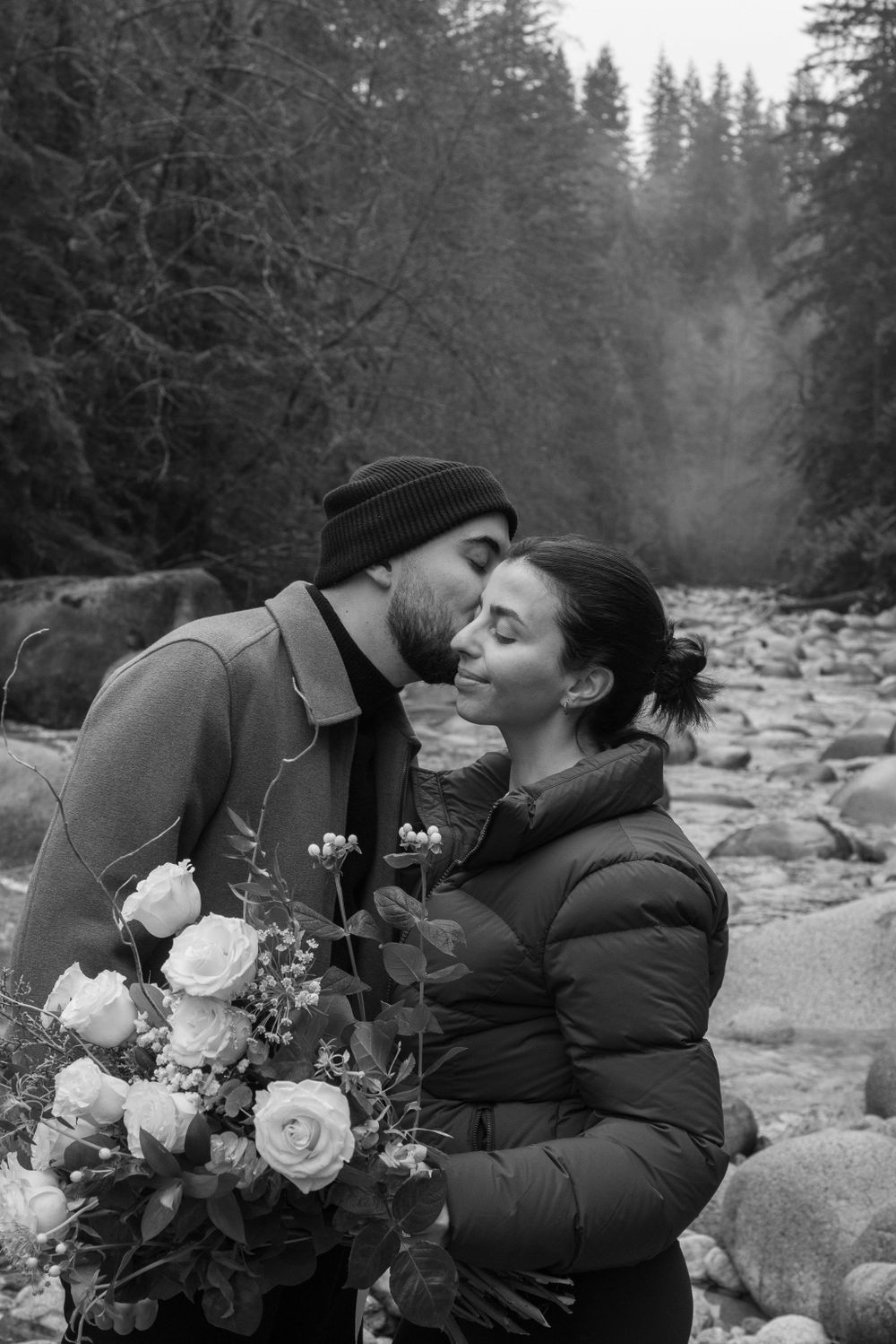 Couple sharing a romantic moment with white roses by evergreen trees in winter.