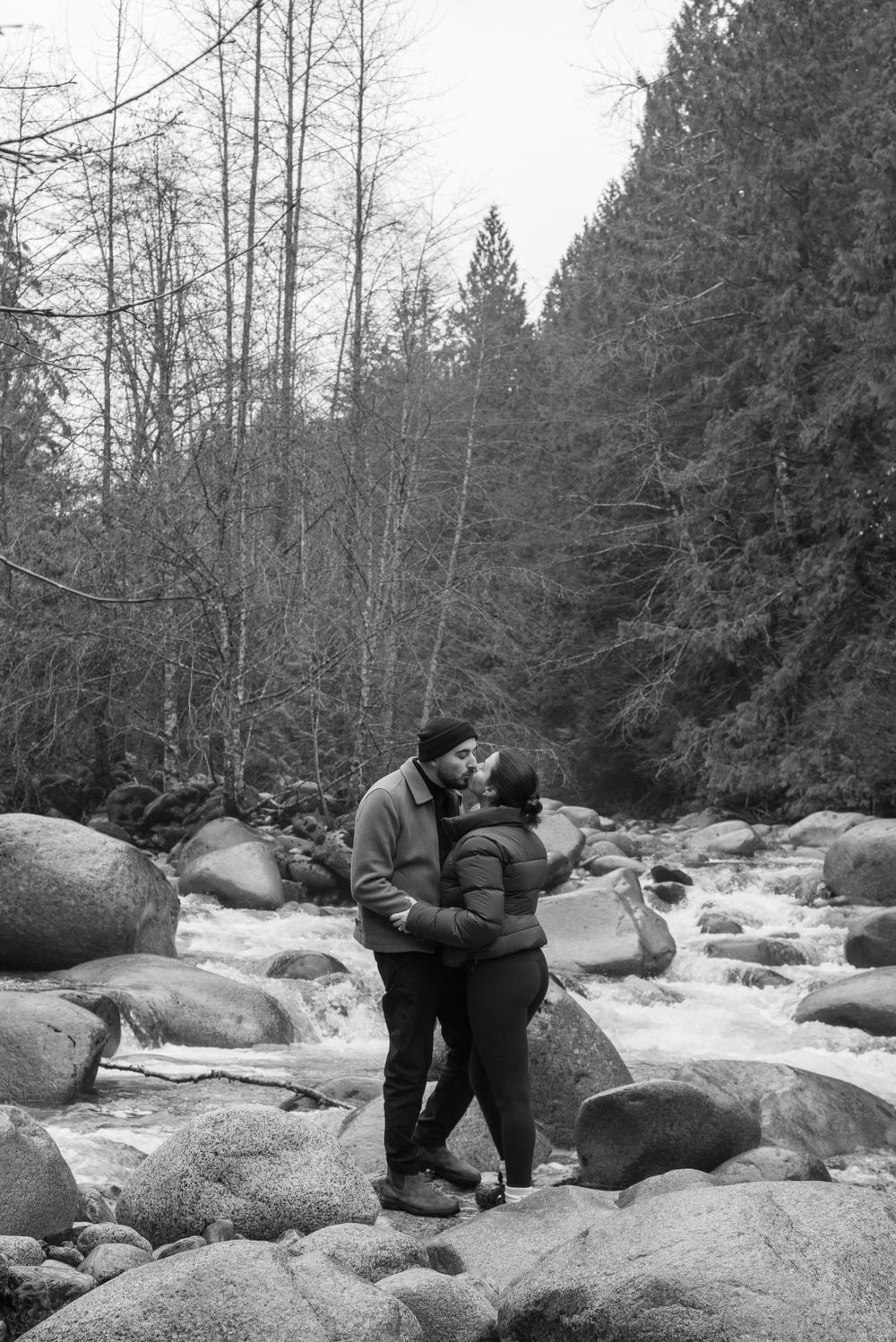 Couple embracing near rushing water and large rocks in a forest setting.