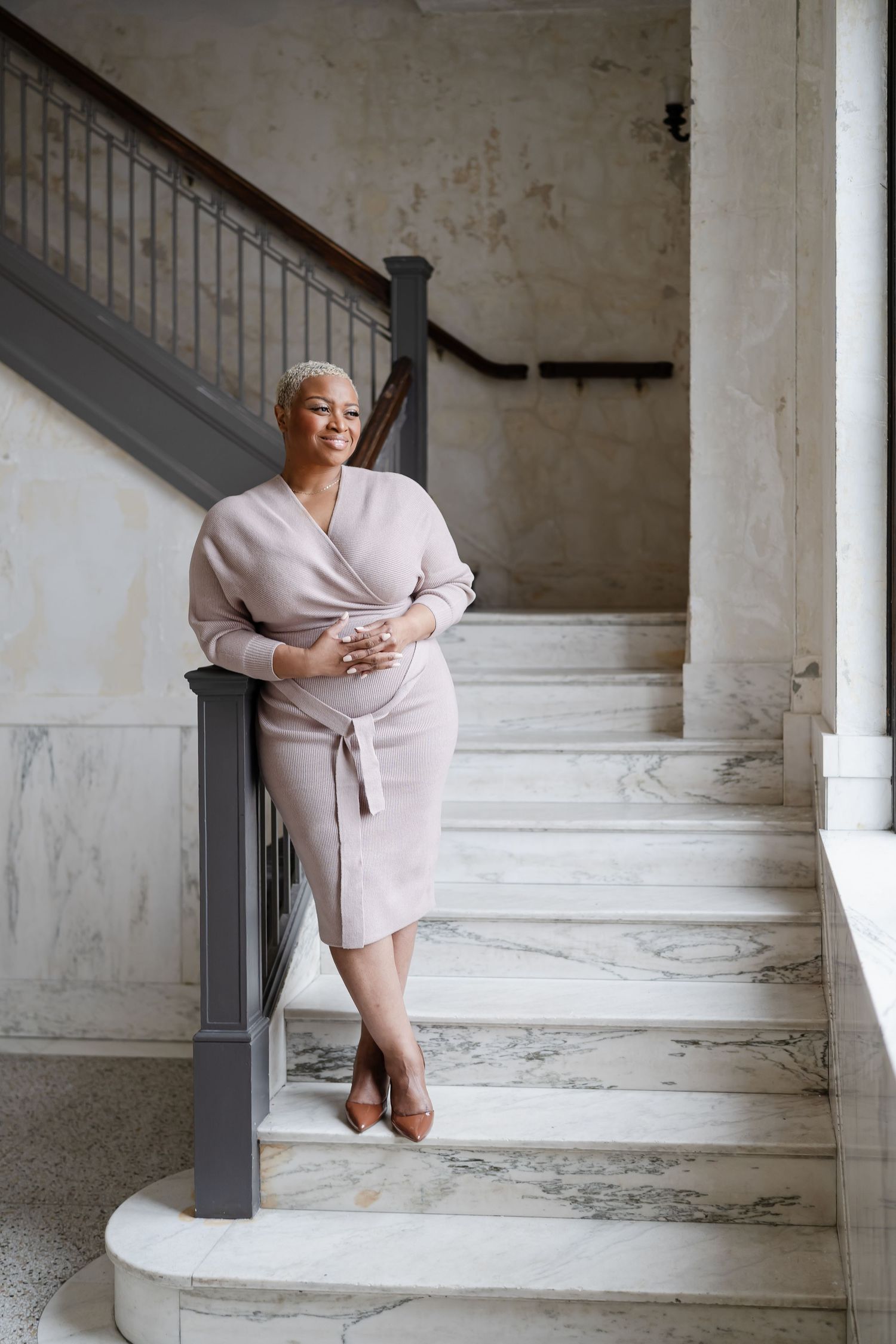 A person in a beige wrap dress and heels poses elegantly on marble stairs in a minimalist interior setting.