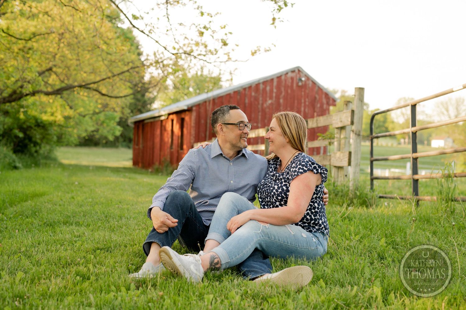 A couple sits together on grass near a red barn and wooden fence on a sunny afternoon.