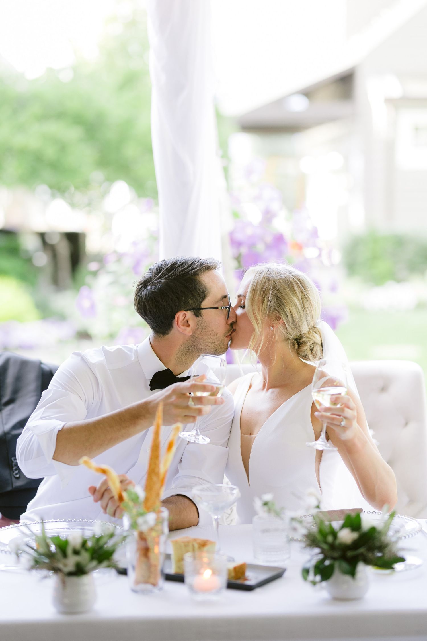 A couple shares a romantic kiss while holding champagne glasses at an elegant table decorated with flowers and candles.