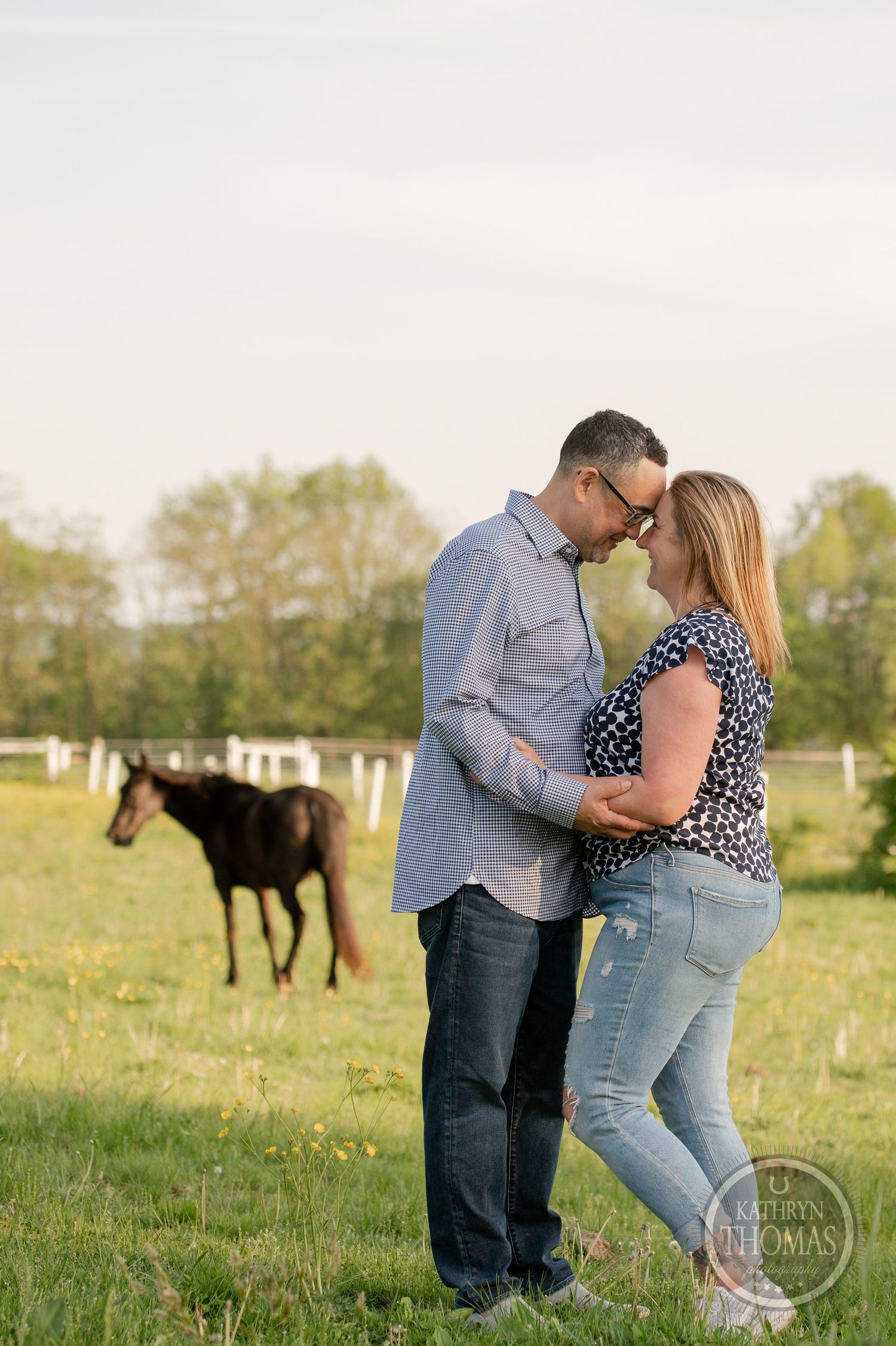 A romantic couple embraces in a field while their horse grazes in the background at sunset.