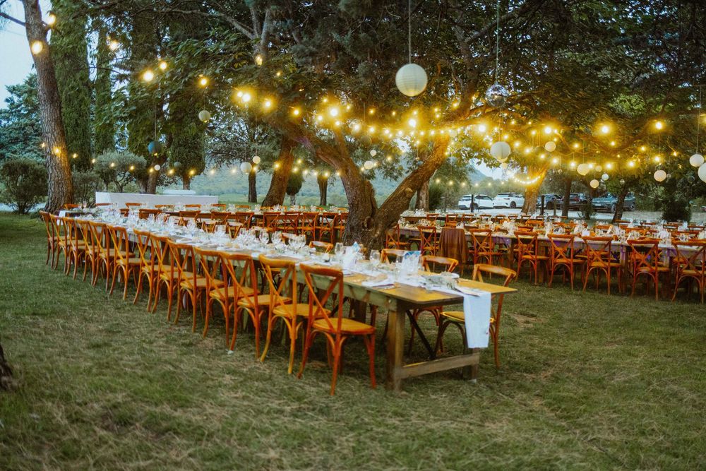 Rustic outdoor wedding reception setup with wooden tables and chairs under string lights at dusk.