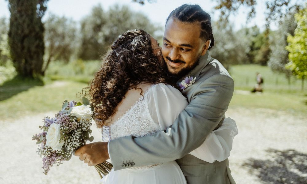 A joyful newlywed couple embraces outdoors, bride holding a floral bouquet in a sunlit park setting.