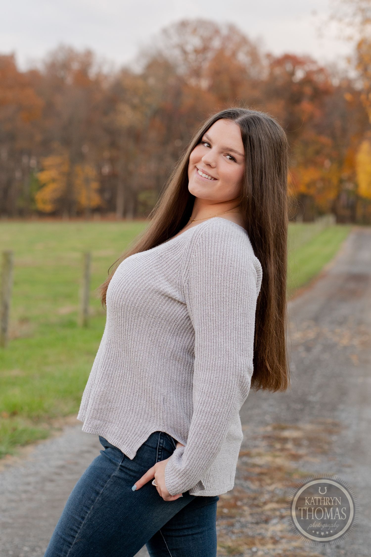 A person poses on a country path wearing a light sweater against a backdrop of fall foliage.