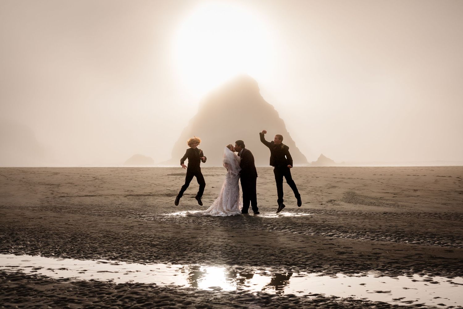 Wedding party jumping joyfully on misty beach at sunset with iconic Haystack Rock silhouetted in background.