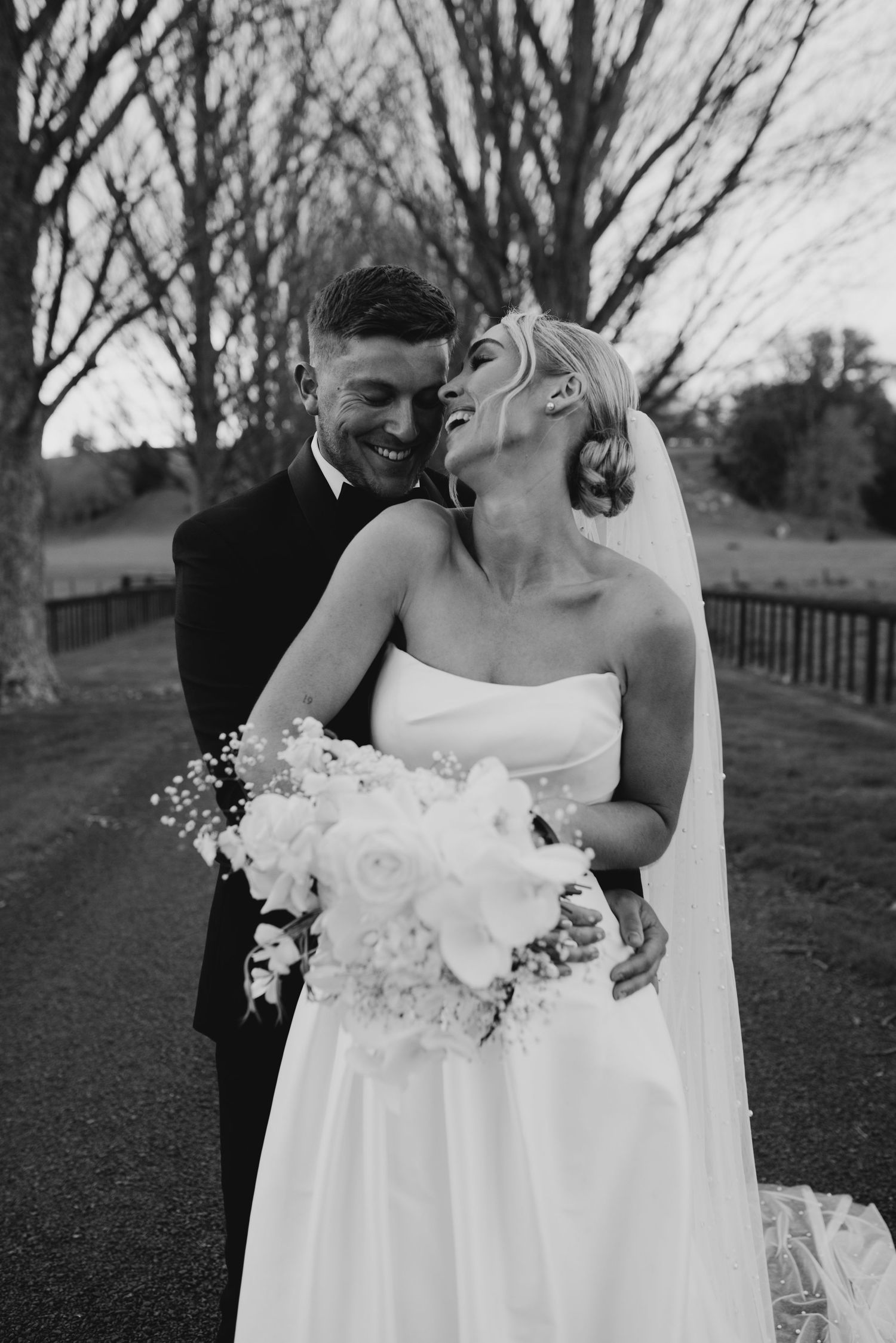 Smiling bride and groom embrace on a tree-lined path holding a white bouquet in black and white.