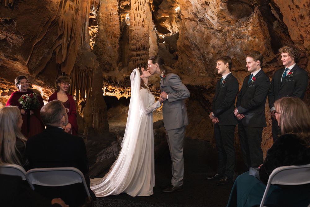 Bride and groom share first kiss during romantic underground cave wedding ceremony with stalactites and wedding guests.