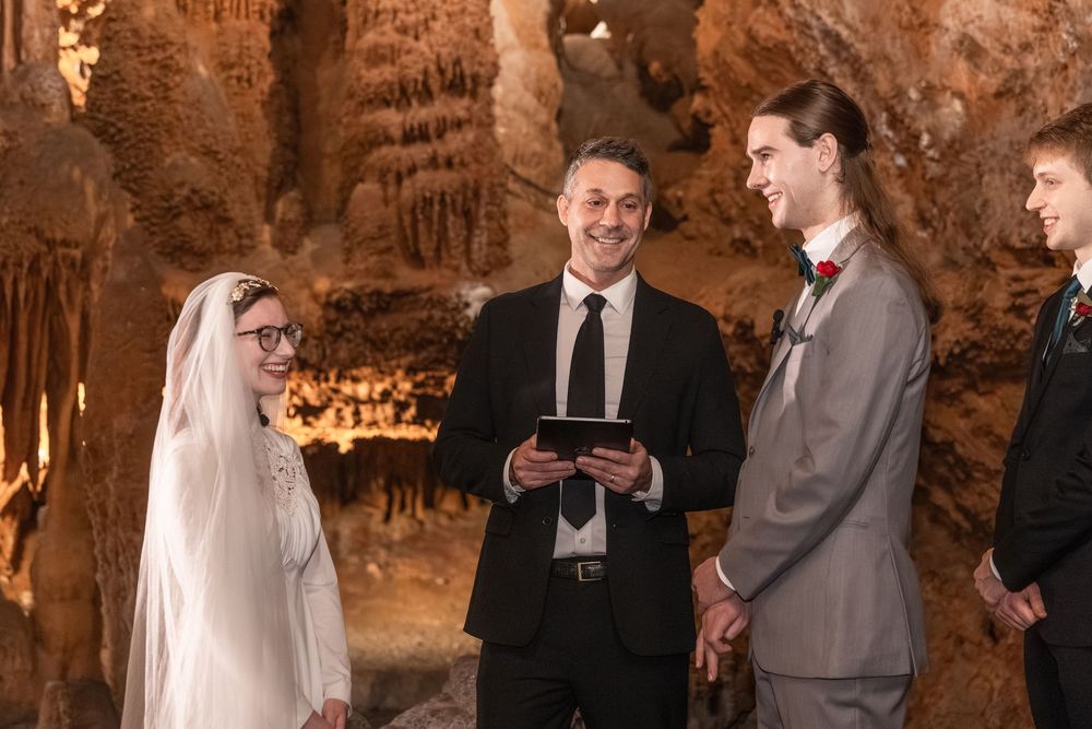 Couple exchanging wedding vows in a stunning cave ceremony with stalactites, officiant holding tablet, bride in veil.