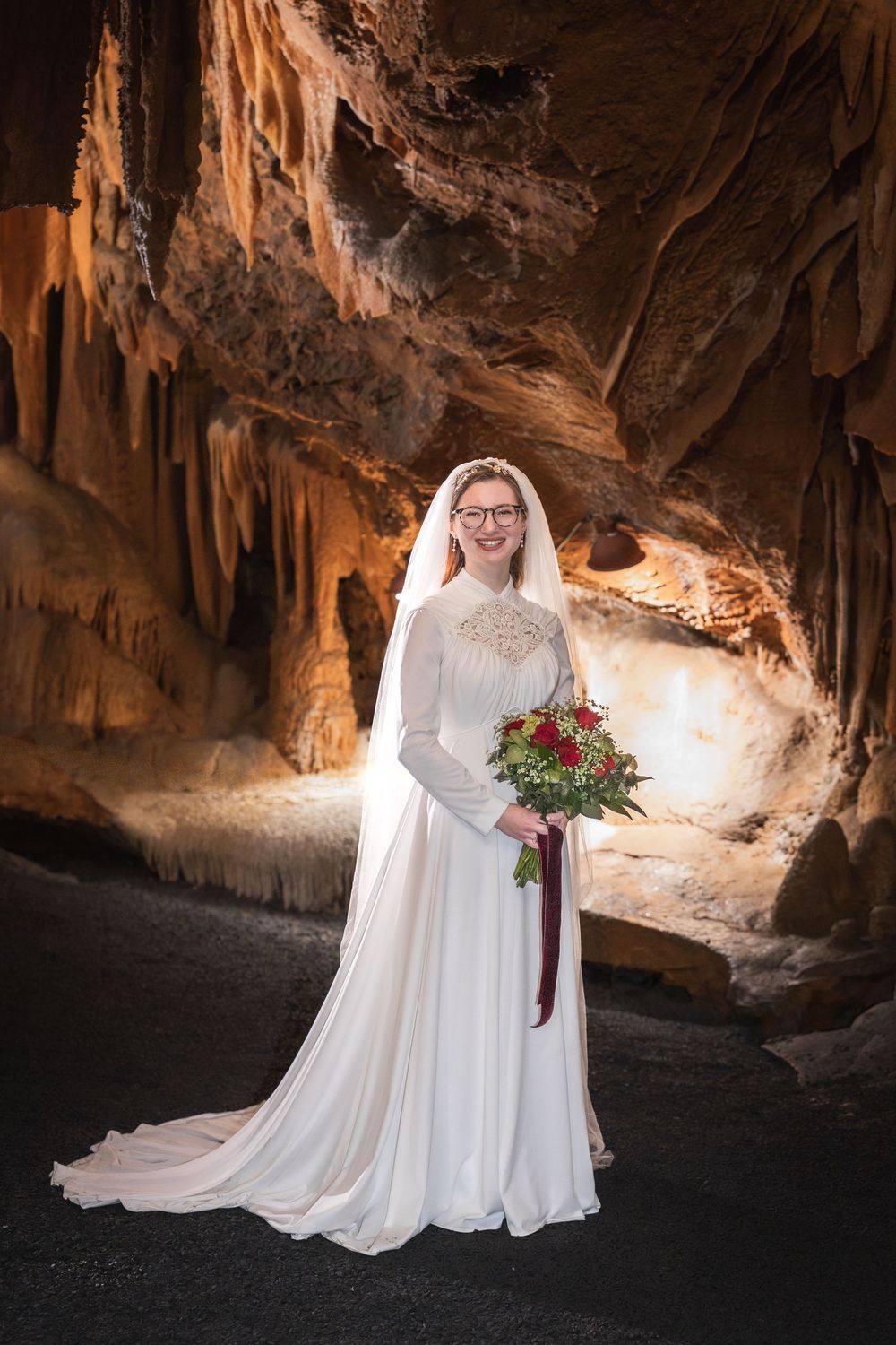 Bride in white gown and veil holding red bouquet, posing inside a stunning cave with stalactites.