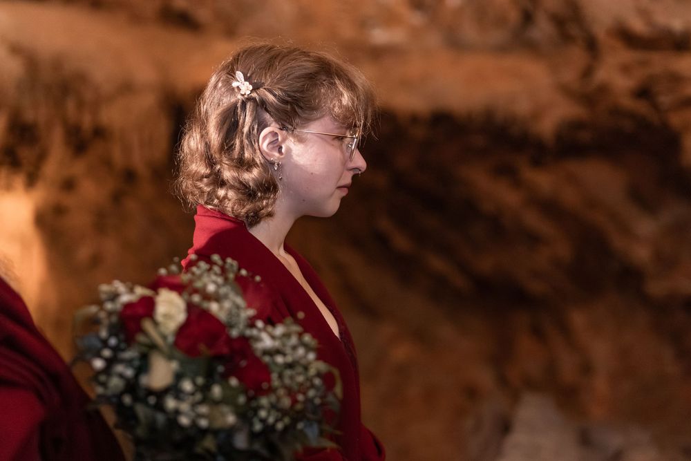 Young woman in red dress holding bouquet of red roses and white flowers in a cave wedding ceremony.