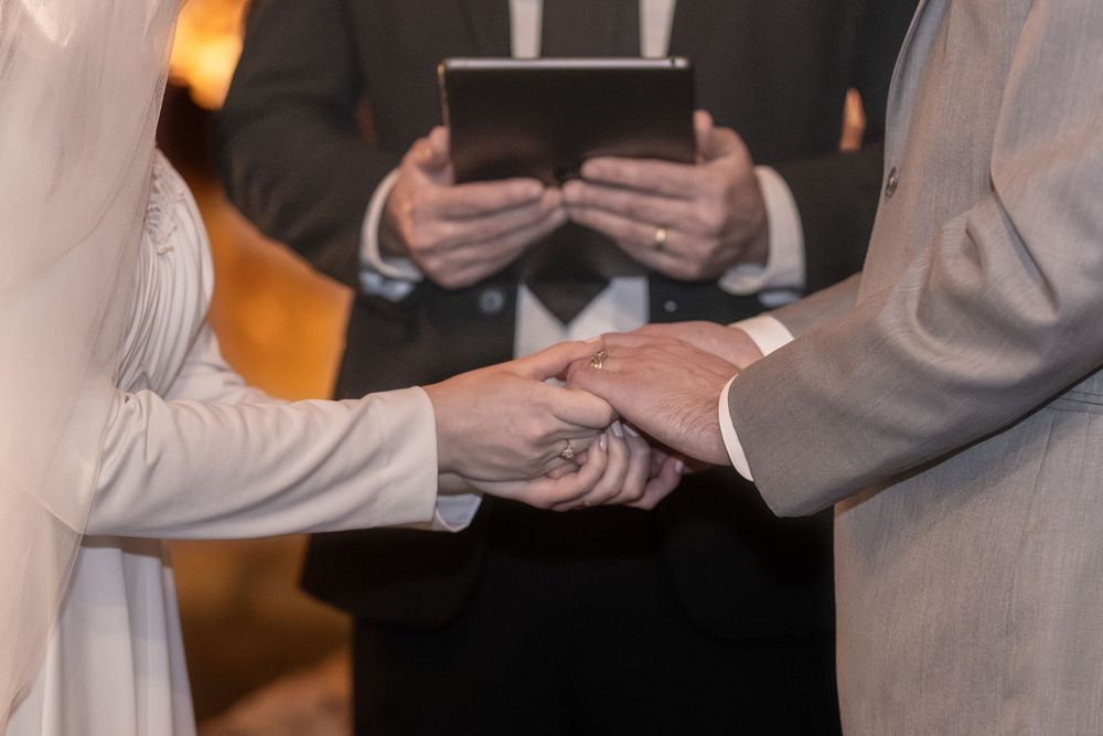 Bride and groom holding hands during wedding ceremony with officiant holding tablet in background.