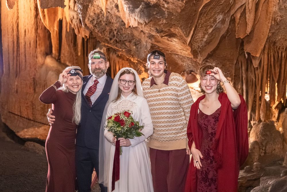 Five people pose for a wedding photo inside a stunning cave, bride holding red roses bouquet.