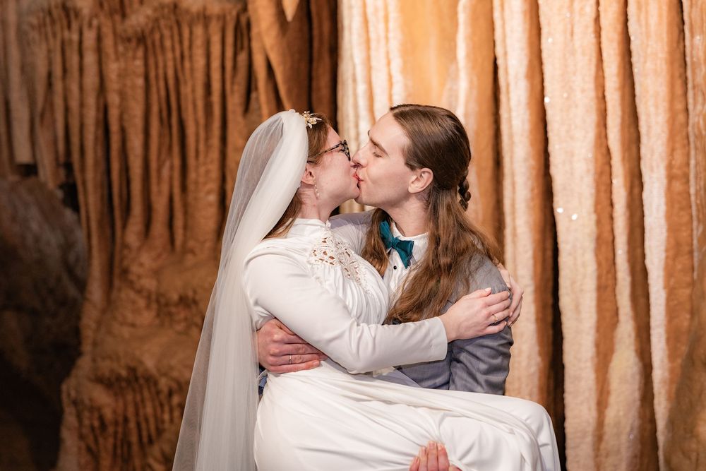 Two brides share a kiss in a cave wedding venue, one wearing a white gown and veil, the other in a gray suit.