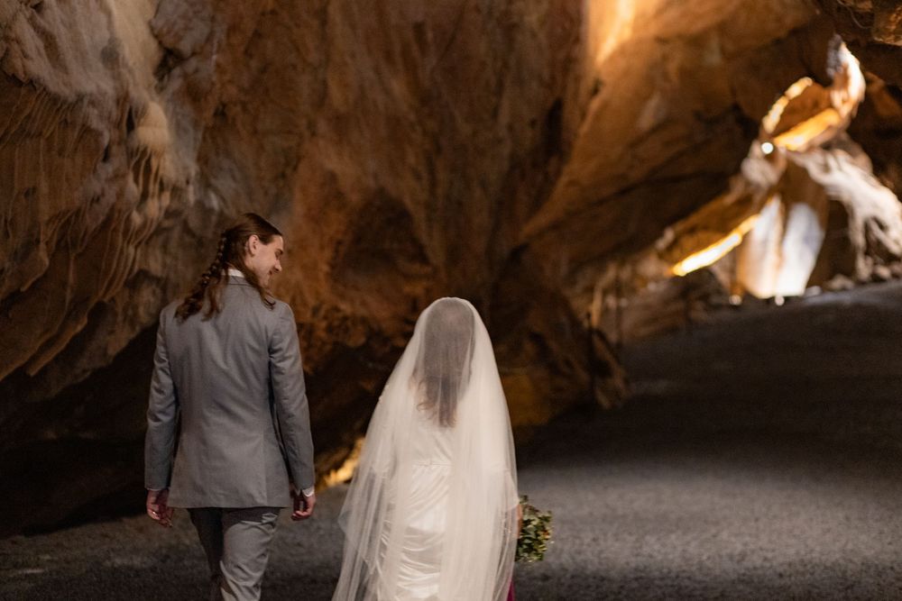 A bride and groom walk hand in hand through a dramatic cave tunnel, her veil trailing behind them.