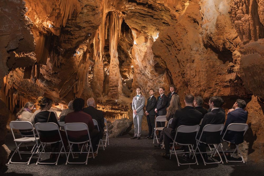 A wedding ceremony held inside a stunning cave with stalactites, guests seated in folding chairs facing the couple.