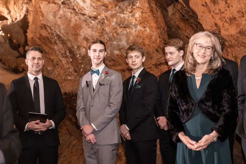 Group of five people posing in a cave wedding venue, with groom in gray suit and boutonniere.