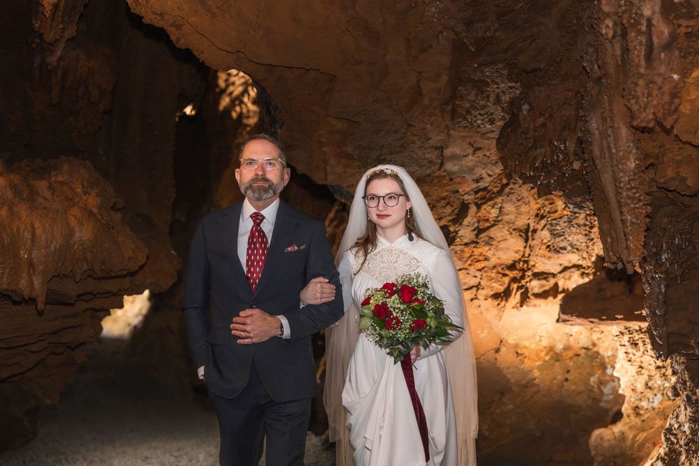Bride and groom walking through a stunning cave wedding venue, bride holding red rose bouquet wearing veil and white gown.