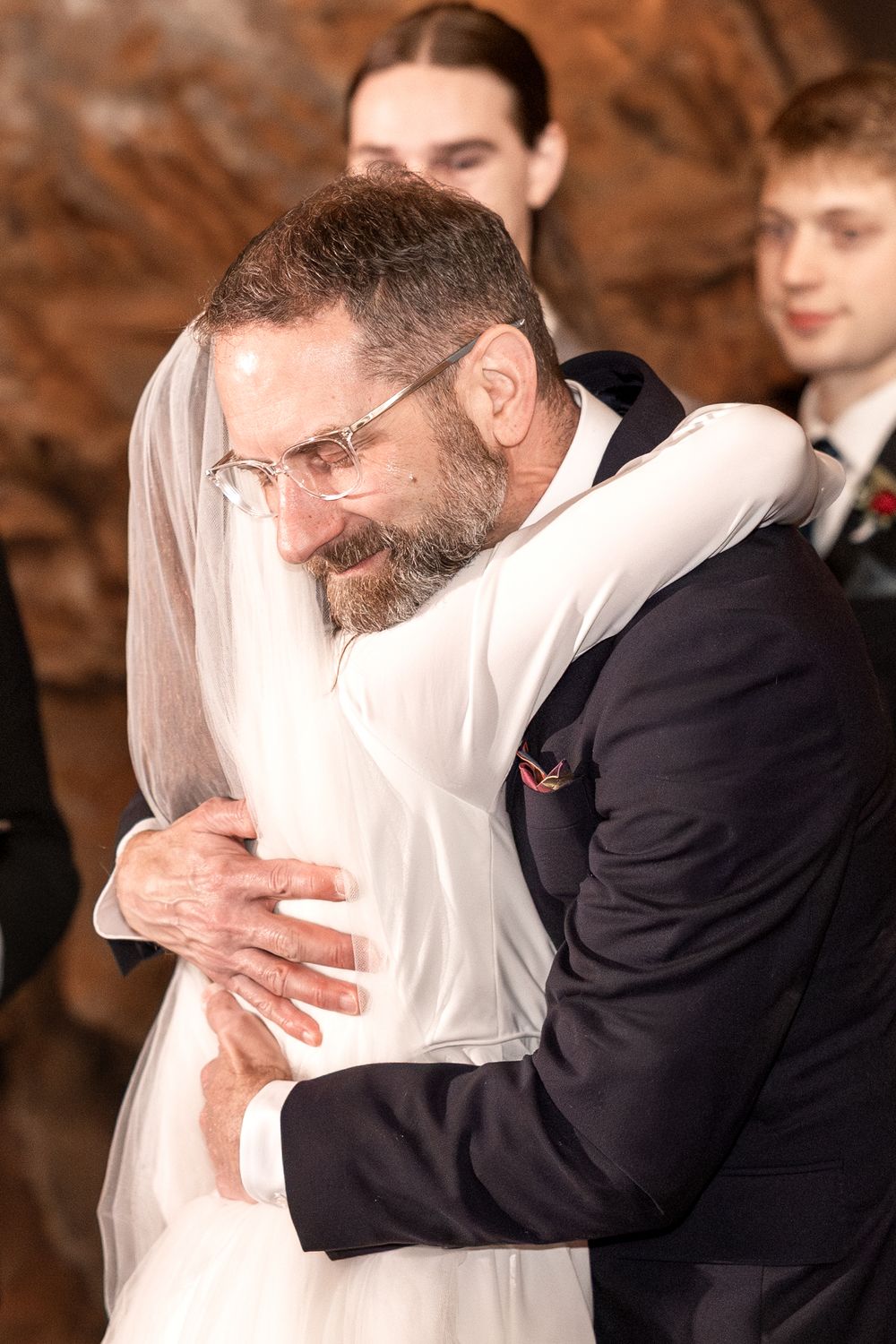 Emotional father in suit hugging bride in white wedding dress and veil during wedding ceremony.