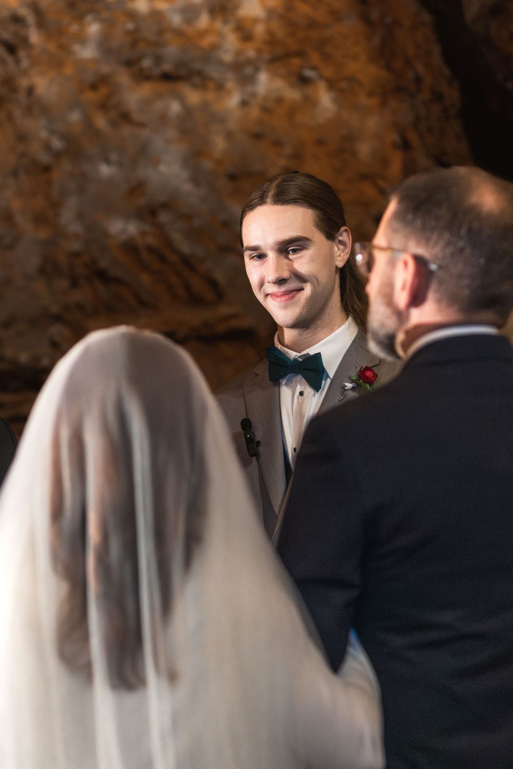 Groom smiling at bride during wedding ceremony in a cave venue, wearing tuxedo with green bow tie and rose boutonniere.