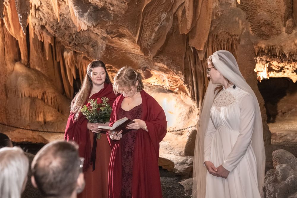 A fantasy-themed cave wedding ceremony with two women in red gowns and a bride in white amid stunning stalactite formations.