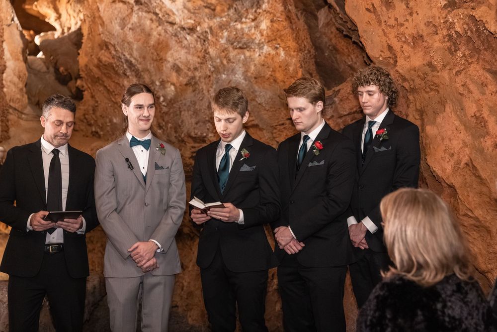 Five men in formal suits stand during a wedding ceremony inside a stunning natural cave with rocky walls.