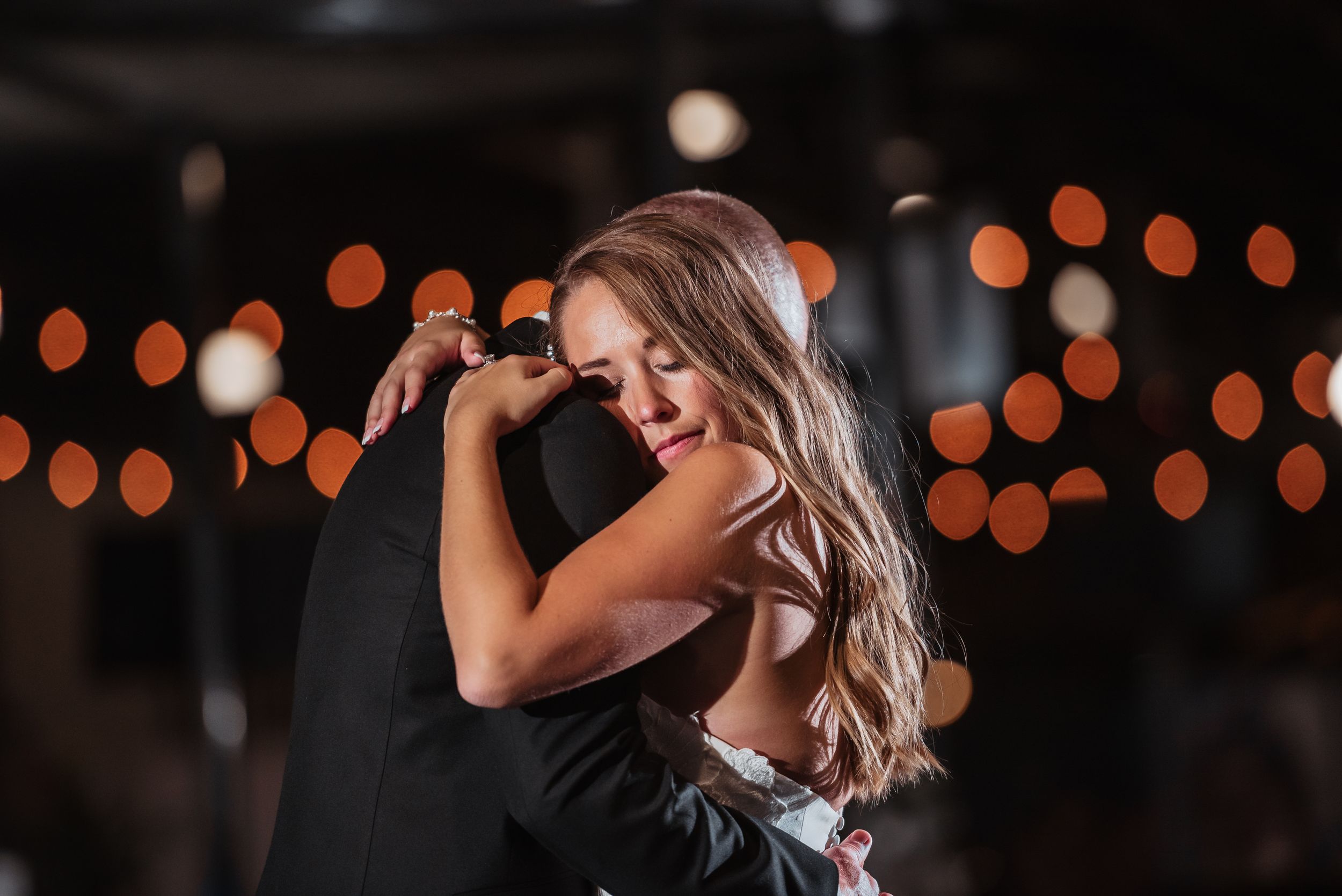 Couple sharing an emotional first dance embrace at a romantic wedding reception with warm bokeh lights in the background.