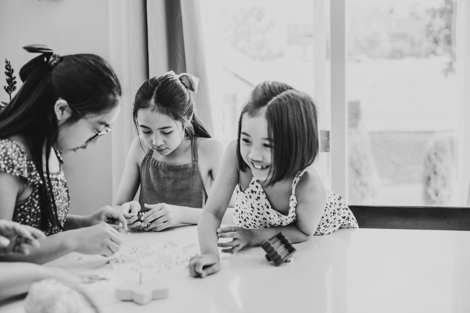 3 girls who are sisters sit at a kitchen counter concentrate and smile while making cookies.