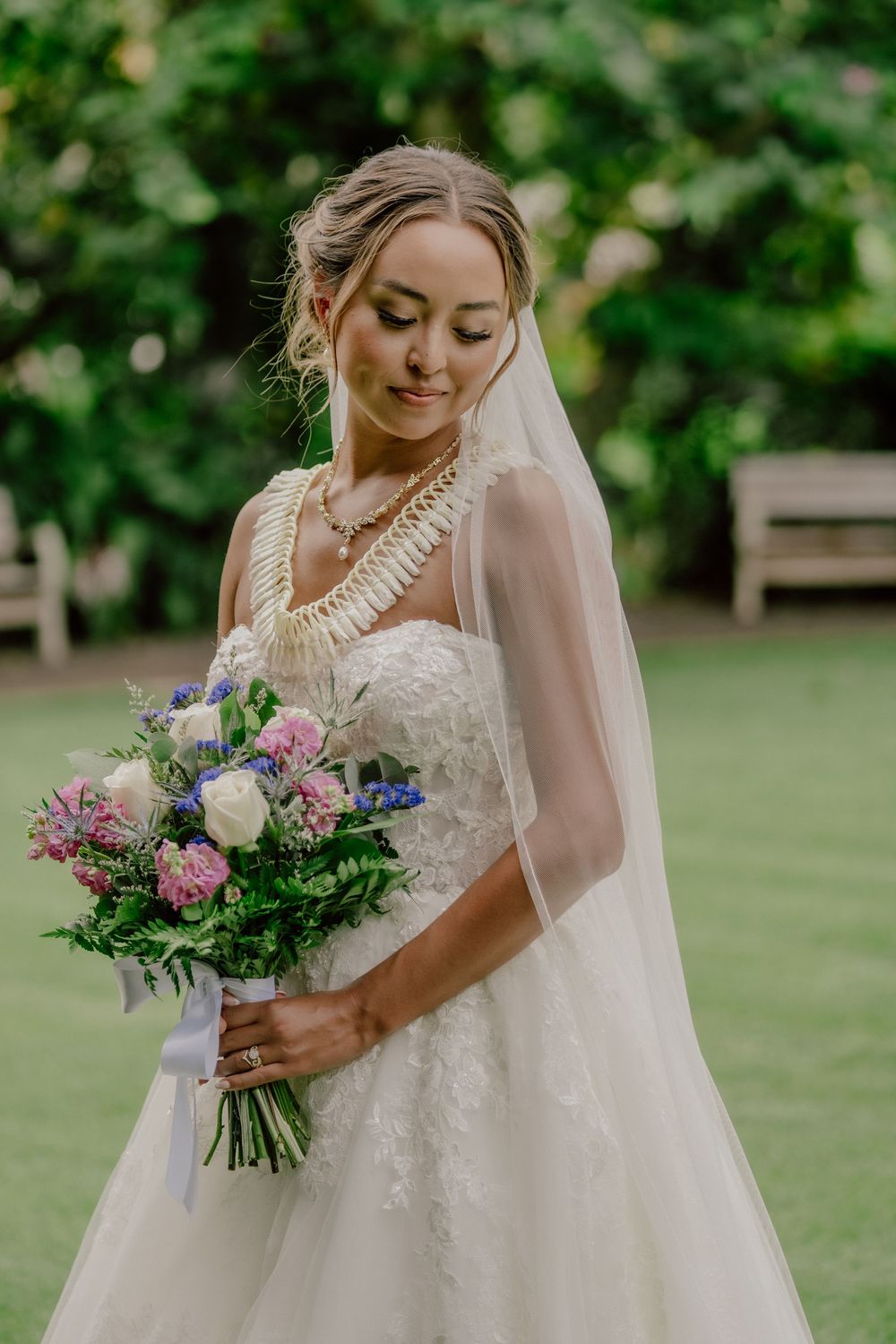 Bride in lace gown and veil holds colorful bouquet of pink, white, and blue flowers outdoors.