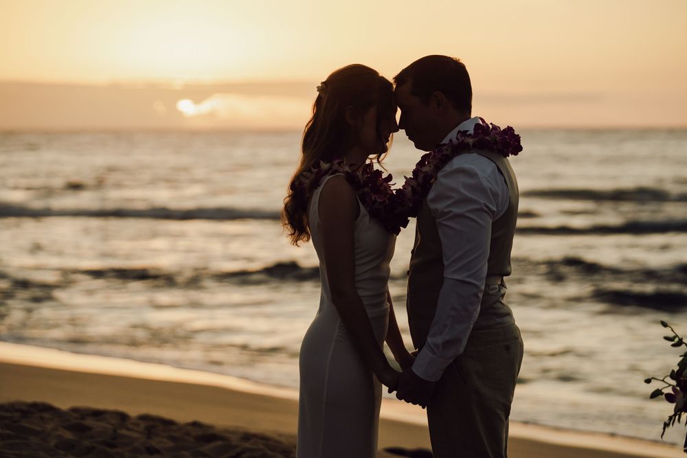 Couple in silhouette sharing an intimate moment on a beach at sunset, wearing floral leis in a romantic wedding scene.