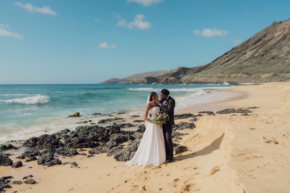 Bride and groom stand on a sandy beach with turquoise ocean waves and rocky shoreline under a bright blue sky.