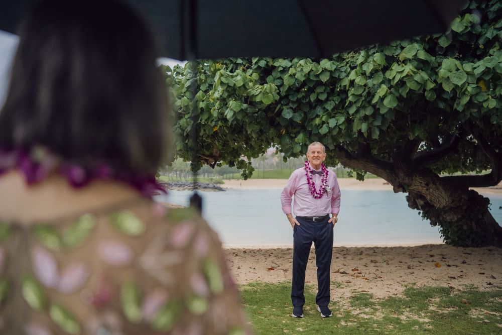 Man wearing lei stands under tropical tree by beach in rain, woman with umbrella in foreground.