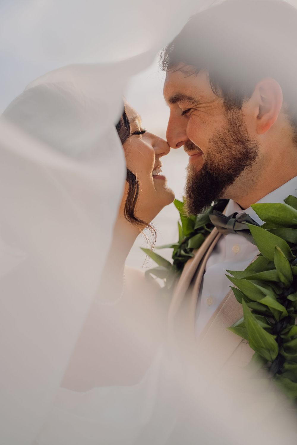 A smiling bride and bearded groom share a romantic moment under a flowing white veil, adorned with tropical green lei.