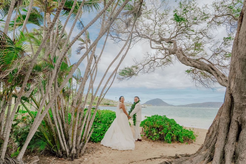Bride and groom stand under tropical trees on a sandy Hawaii beach with turquoise ocean views on their wedding day.