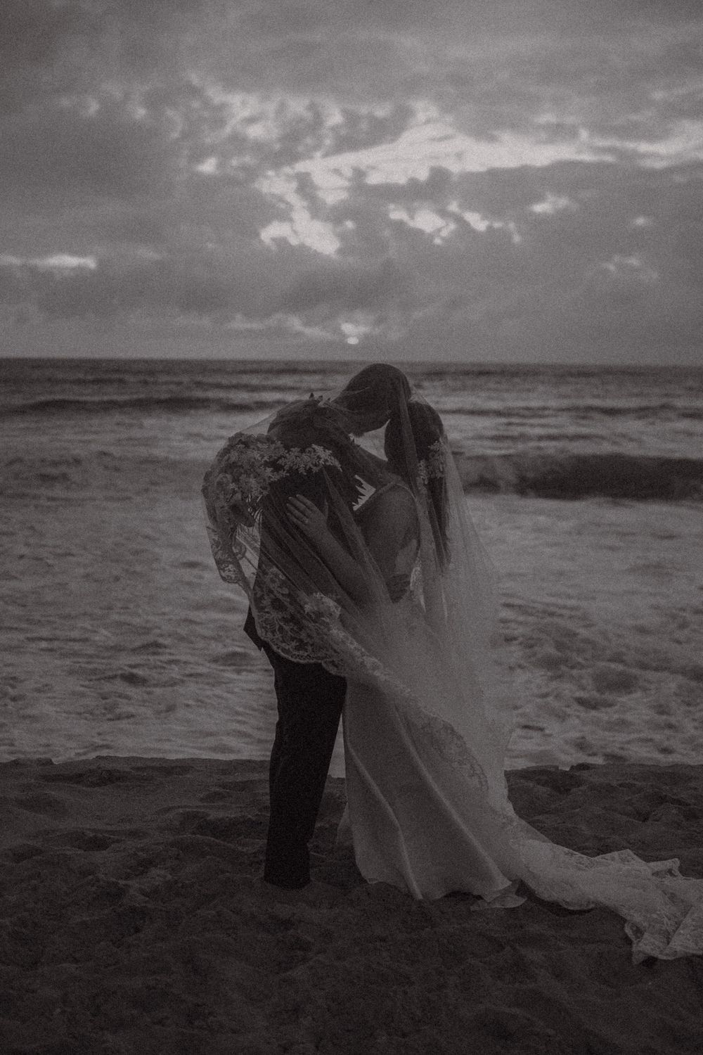 Black and white romantic photo of newlyweds embracing on beach at dusk with dramatic cloudy sky.